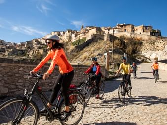 Gruppe von Radfahrern auf einem Kopfsteinpflasterweg in der Region Valencia mit einem historischen Bergdorf und Festungsmauern im Hintergrund.