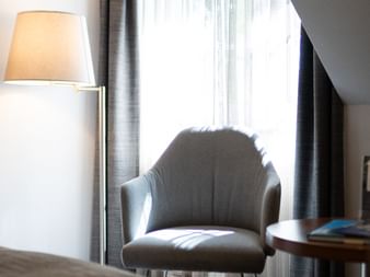 Grauer gepolsterter Sessel mit Fellkissen neben einem Fenster mit Gardinen und grauen Vorhängen. Eine Stehlampe und ein kleiner Beistelltisch.