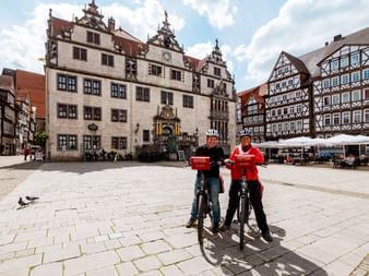 Zwei Radfahrer mit Rädern auf dem Marktplatz in Hann. Münden. Historisches Rathaus und Fachwerkhäuser unter blauem Himmel mit Wolken.