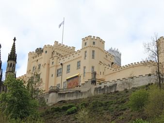 Schloss Stolzenfels auf einem Hügel bei Koblenz mit gelben Mauern, Türmen und Zinnen. Links ist eine gotische Kirchturmspitze sichtbar.