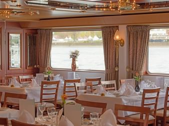 Eleganter Speisesaal auf der MS Switzerland mit gedeckten Tischen, Holzstühlen, weißen Tischdecken und großen Fenstern mit Flussblick.