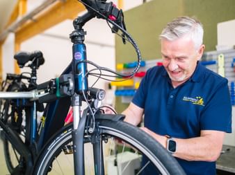 Älterer Mann mit grauen Haaren in blauem Poloshirt arbeitet an schwarzem Fahrrad in professioneller Reparaturwerkstatt mit Werkzeugen.