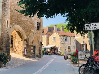 Radfahrer mit Fahrrad in einem mittelalterlichen Steindorf in der Dordogne, Frankreich. Steinbogen, traditionelle Häuser und Straßenschild unter Baumschatten.
