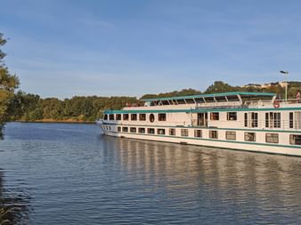 Flusskreuzfahrtschiff MS Switzerland mit weißem Rumpf und grünen Akzenten auf ruhigem Fluss, umgeben von Bäumen unter blauem Himmel.