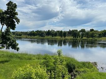 Ruhiger Fluss Memel im Baltikum mit grünen grasbewachsenen Ufern, Bäumen und bewölktem blauen Himmel, der sich im Wasser spiegelt.