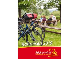 Cover des Rückenwind Radreisen Katalogs 2019 mit Fahrrädern mit roten Taschen auf Wiese und Menschen an einem Picknicktisch unter Bäumen.
