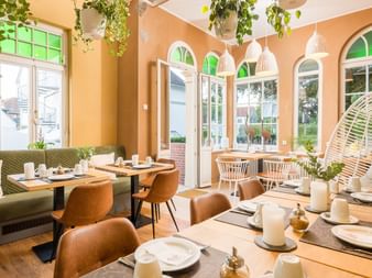 Helles Restaurant mit pfirsichfarbenen Wänden, Holztischen, braunen Stühlen, hängenden Pflanzen und Bogenfenstern. Tische sind mit weißen Tellern gedeckt.