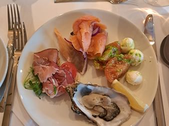 Eleganter Vorspeisenteller mit geräuchertem Lachs, Schinken, frischer Auster, Cherrytomate, Mozzarellakugeln und Zitronenschnitz auf weißem Porzellan.