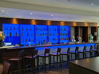 Zeitgenössische Hotelbar im Sheraton Essen mit blau beleuchteten Regalen voller Flaschen, dunkler Bartheke und Reihe moderner Barhocker.