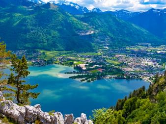 Panoramablick auf den Traunsee im Salzkammergut von erhöhtem Felsaussichtspunkt mit türkisem Wasser, umliegender Stadt und schneebedeckten Alpen.