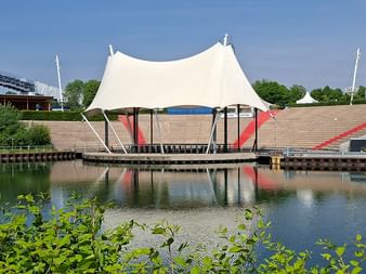 Amphitheater Gelsenkirchen mit weißem zeltartigem Dach über schwimmender Bühne, umgeben von Wasser und Sitzreihen unter blauem Himmel.