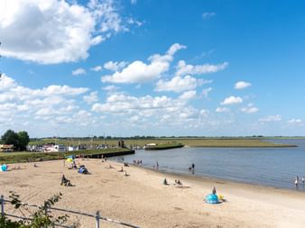Sandstrand in Dangast mit vereinzelten Besuchern, hölzerner Steg ins ruhige Wasser, blauer Himmel mit weißen Wolken und grünen Bäumen links.