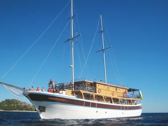 Weißes Segelschiff San Snova mit zwei Masten und Holzdeck auf blauem Wasser vor kroatischer Küste unter klarem Himmel.