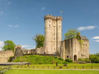 Mittelalterliche Burgruine Kasselburg mit Doppelturm und Steinmauern auf grünem Hügel, umgeben von Holzzaun unter blauem Himmel.