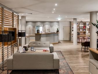 Moderne Hotellobby mit grauen Sofas, Holzraumteiler, Rezeptionstresen und Zierpflanzen. Heller Holzboden im gesamten Raum.