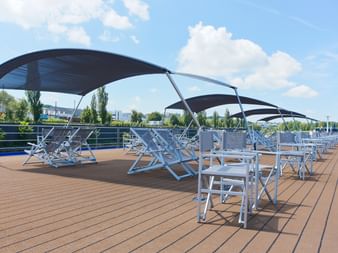 Sonnendeck mit Holzboden mit blau-weißen Klappliegen unter schwarzen Sonnendächern. Metallgeländer säumen das Deck, Bäume im Hintergrund sichtbar.
