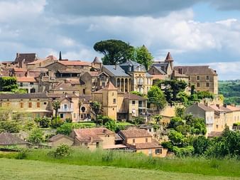 Dorf am Hang in der Dordogne mit Steingebäuden, Kirchturm und Terrakottadächern, umgeben von grünen Feldern und Bäumen unter bewölktem Himmel.