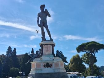 Bronzekopie von Michelangelos David-Statue auf verziertem Sockel auf toskanischem Platz, umgeben von parkenden Autos und Mittelmeerkiefern.