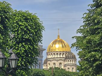 Goldene Kuppel der Christi-Geburt-Kathedrale in Riga, eingerahmt von grünen Bäumen. Die byzantinische Kuppel glänzt unter blauem Himmel.