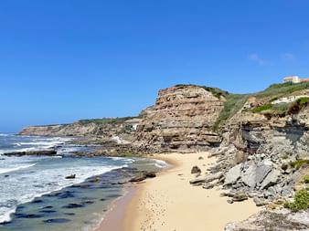 Küstenansicht der Bucht São Pedro da Cadeira mit geschichteten Sandsteinklippen, Sandstrand und Atlantikwellen unter blauem Himmel.