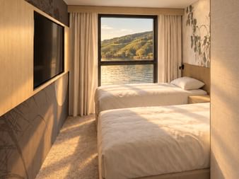 Modern twin cabin on MS Veloria with two beds, wooden paneling, wall-mounted TV, and large window showing river and hillside landscape.