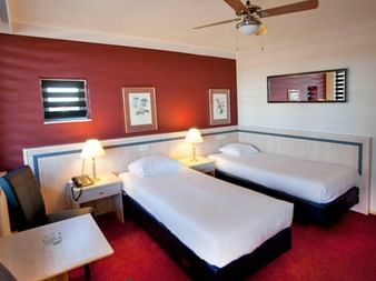 Modernes Hotel-Zweibettzimmer mit zwei Einzelbetten, roter Akzentwand, weißen Möbeln, Schreibtisch mit Stuhl und Deckenventilator.