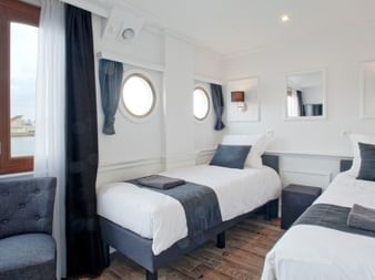 Modern ship cabin on MS Magnifique II with two single beds, round portholes, blue curtains, and a chair by the window overlooking water.