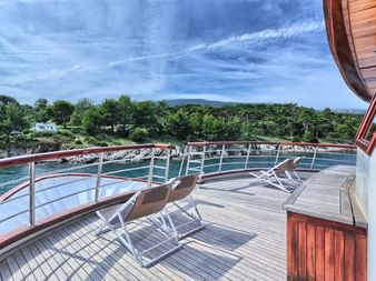 Bug-Sonnendeck der MY Andela Lora mit Holzdeck, Liegestühlen, Metallgeländer und Blick auf türkisfarbenes Wasser und grüne Küste.