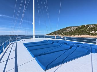 Sonnendeck des Schiffes San Snova mit blauen gepolsterten Liegen, weißem Mast und Reling. Kroatische Küste mit Hügeln im Hintergrund.