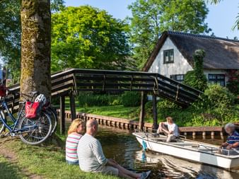 Menschen entspannen an einem Kanal in Giethoorn, während ein Motorboot unter einer Holzbrücke vorbeifährt. Zwei Fahrräder stehen unter einem Baum neben einem Reetdachhaus.