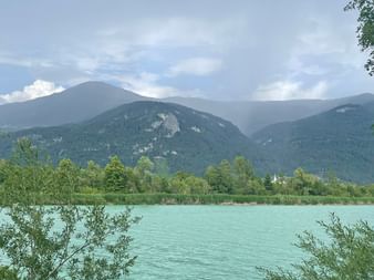 Türkisfarbener See umrahmt von grüner Vegetation mit bewaldeten Bergen im Hintergrund unter teilweise bewölktem Himmel in der Alpe-Adria-Region.