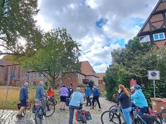 Große Gruppe von Radfahrern mit Fahrrädern versammelt auf Kopfsteinpflasterplatz umgeben von traditionellen roten Backsteingebäuden mit Ziegeldächern.