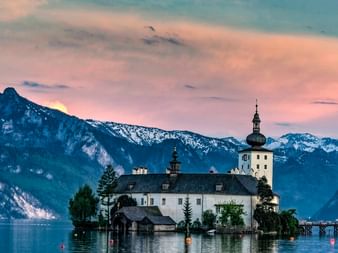 Schloss Orth auf einer Insel im Traunsee mit schneebedeckten Bergen im Hintergrund unter rosa-blauem Abendhimmel im Salzkammergut.