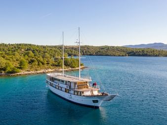 Weiße Motoryacht MY Magellan mit zwei Masten fährt durch türkisfarbenes Wasser entlang der bewaldeten kroatischen Küste unter blauem Himmel.