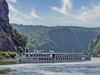 MS Veloria river cruise ship sailing on calm water between steep rocky cliffs covered with green vegetation under a blue sky.
