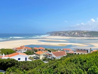 Luftaufnahme des Strandes Praia do Bom Sucesso in Portugal mit weißem Sand, blauen Meereswellen und rotgedeckten Häusern zwischen grüner Vegetation.