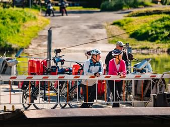 Zwei Radfahrer mit Helmen und Fahrrädern stehen auf einer kleinen Fährplattform in Hemeln. Die Fähre hat rote Ausrüstung und überquert einen Fluss.