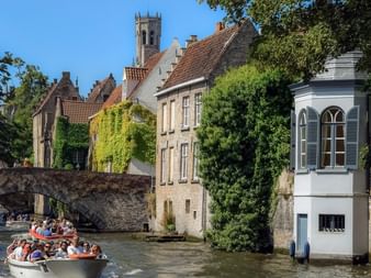 Touristenboote auf einem Kanal in Brügge mit historischen Gebäuden, einer Steinbrücke und einem Kirchturm. Efeubedeckte Fassaden säumen den Wasserweg.