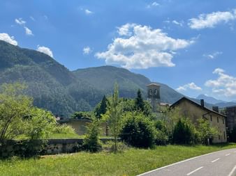 Asphaltierte Straße durch Alpendorf mit Kirchturm, umgeben von grünen Wiesen und bewaldeten Bergen unter blauem Himmel mit Wolken.