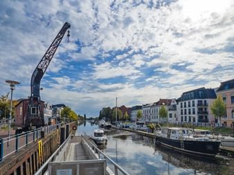 Alter Hafenkran am Wasser in Oldenburg mit Booten am Kanal. Historische Gebäude säumen beide Seiten unter bewölktem Himmel.