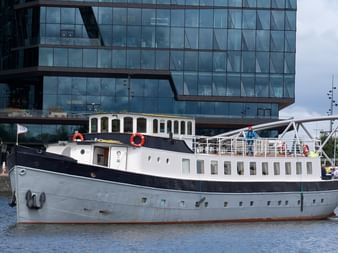 Passagierschiff Floor van Aemstel mit dunkelblauem Rumpf und weißem Aufbau im Hafen vor modernem Glasgebäude.