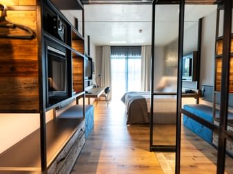 Zeitgenössisches Hotelzimmer mit Glastrennwand, Holzakzenten, TV-Einheit und blauen Sitzgelegenheiten. Bett durch Glaswand sichtbar mit Tageslicht.