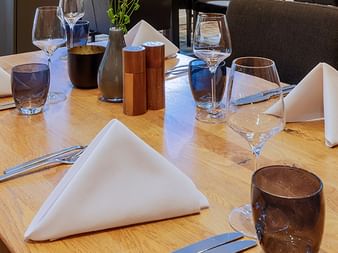 Elegant gedeckte Restauranttische mit weißen Servietten, Weingläsern, Wassergläsern und kupferfarbenen Accessoires auf hellem Holz.