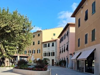 Fußgängerplatz in Casciani Terme mit bunten italienischen Gebäuden in Gelb, Rosa und Beige. Grüne Fensterläden, Markisen und Baum mit Blumenbeet.