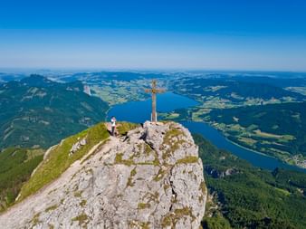 Luftaufnahme des Schafberg-Gipfels mit Holzkreuz und Besuchern auf felsigem Gipfel, Blick auf Wolfgangsee und Salzkammergut-Berge.
