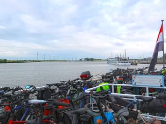 Zahlreiche Fahrräder auf einem Fährdeck mit niederländischer Flagge. Blick über das IJsselmeer mit ferner Küstenlinie unter bewölktem Himmel.