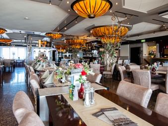 Innenraum des Hotel Old Dutch Restaurants in Volendam mit beigen gepolsterten Stühlen, gedeckten Tischen mit Blumen und orangefarbenen Pendelleuchten.