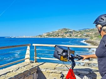 Radfahrerin mit Helm und Rucksack steht mit Fahrrad an Aussichtspunkt und blickt auf blaues Mittelmeer und italienische Küste.