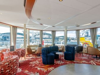Modern ship lounge on MS Lisabelle with curved panoramic windows, blue and orange armchairs, red patterned carpet, and harbor view.