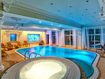 Eleganter Innenpool mit blauer Beleuchtung und Whirlpool im Vordergrund. Klassische Säulen, Kronleuchter und Liegestühle umgeben den Poolbereich.